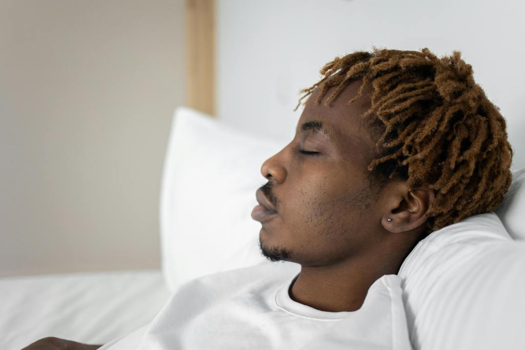 A young man with dreadlocks is peacefully sleeping on a white bed with pillows.