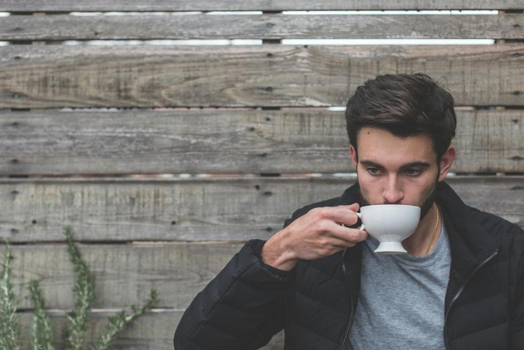 A young man sipping from a white cup against a wooden backdrop.