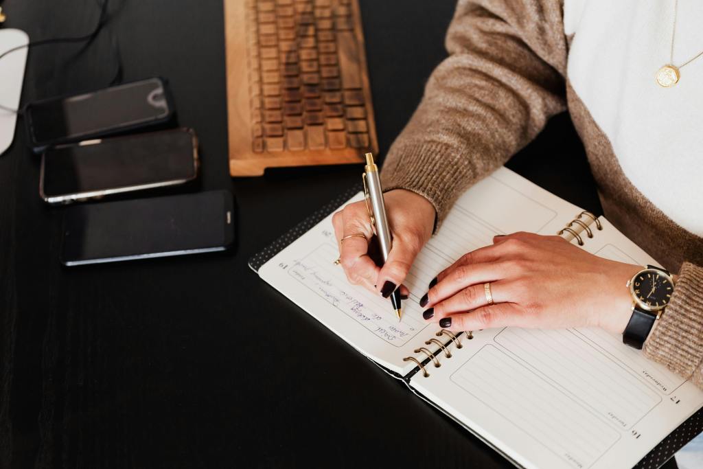 A person writing in a planner with a pen, surrounded by smartphones and a wooden keyboard on a desk.