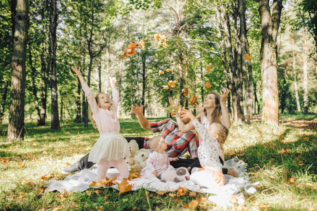 A joyful family with two girls and a baby playing in a forest, throwing autumn leaves in the air while sitting on a blanket with plush toys.